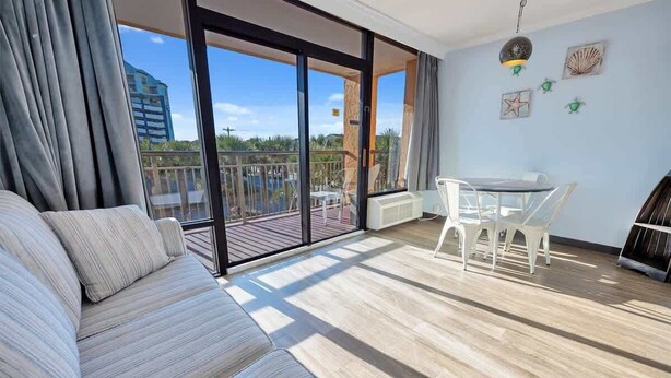A modern living room with a sofa, a dining table with chairs, and a balcony with a view of the city. - Newly Remodeled Caravelle-Studio, Views with Pools (Myrtle Beach)