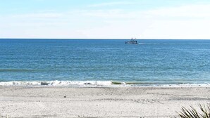 Una playa con un barco a lo lejos, cielo despejado y mar tranquilo.