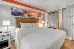 Une chambre d’hôtel avec un grand lit, deux lampes de chevet, une table de chevet, un tableau au-dessus du lit, une porte-fenêtre coulissante et une porte blanche.