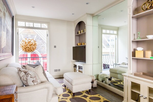 A spacious living room with a sofa, ottoman, and a dog. There is a fireplace, a television, and a bookshelf.