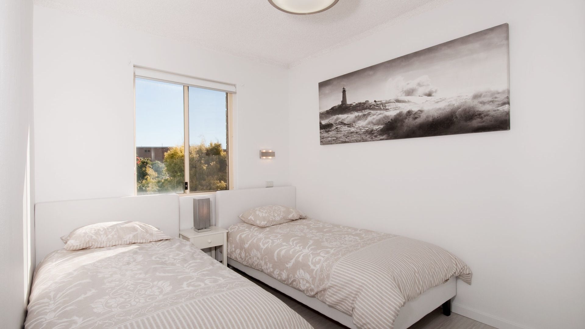 A bedroom with two beds, a window, a wall-mounted light, and a painting of a lighthouse.