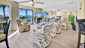 A spacious living room with a large window, a ceiling fan, and a view of the beach. - ☀️Beach Front-Pool + HotTub-Latitude 29 #2☀️ (Panama City Beach)