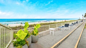 Une promenade en bord de mer, agrémentée de chaises longues et de plantes en pot, qui offre une vue sur une plage de sable où se trouvent des personnes et des parasols.