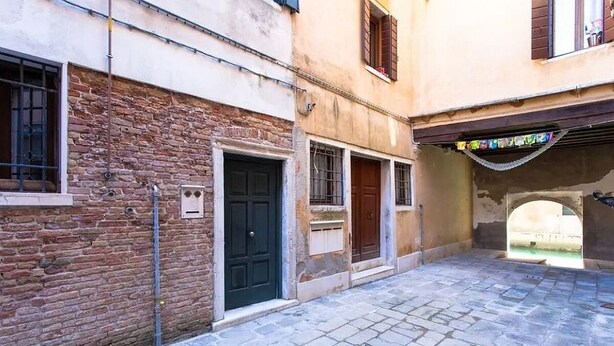 A narrow cobblestone street with buildings on either side, featuring a mix of brick and plaster facades, a black door, and a small window with shutters. - Signorile su Corte Veneziana (Venezia)