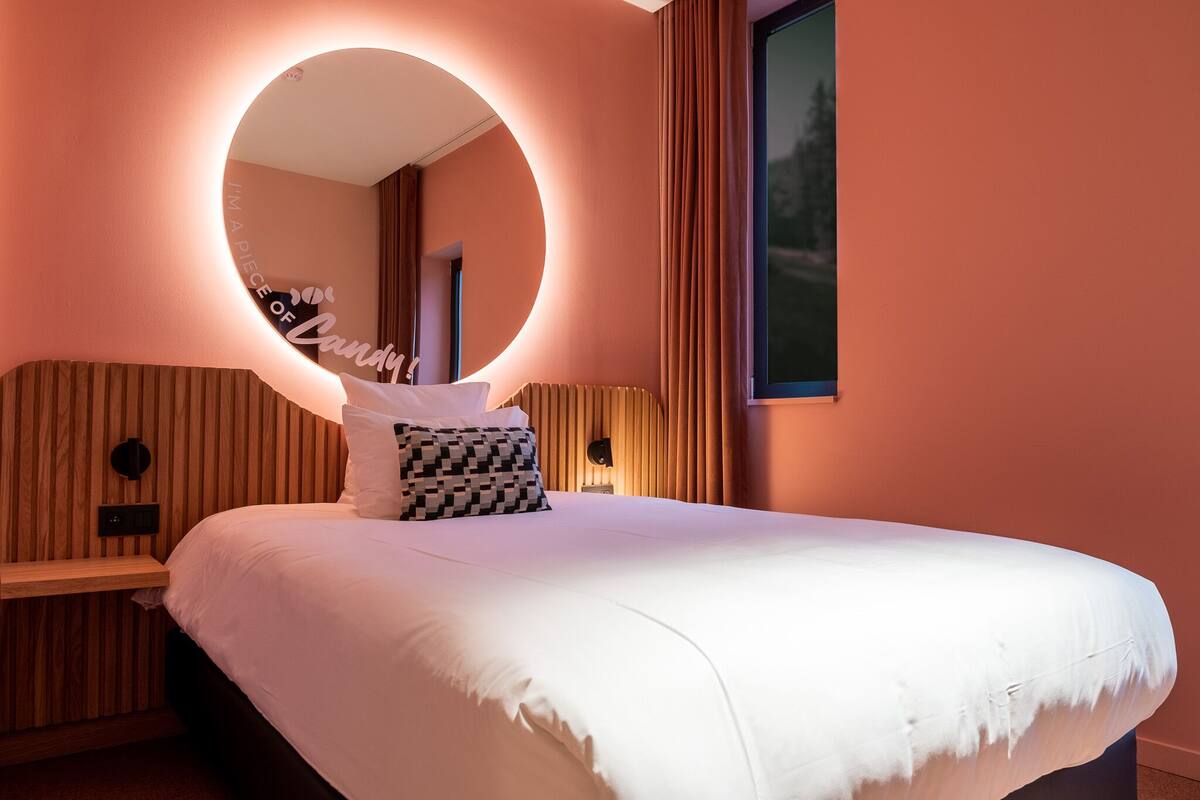 Una habitación de hotel con una cama grande, un espejo redondo, un cabecero de madera y una mesita de noche.