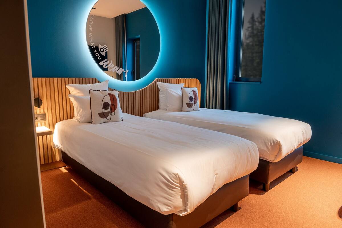 Habitación de hotel con dos camas, un espejo circular y vistas a árboles a través de una ventana.