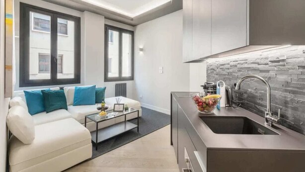 Una sala de estar moderna con un sofá y una mesa de centro, una cocina con salpicadero de piedra y ventanas amplias.