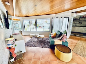 A modern living room with a wooden ceiling, large windows, a green sofa, a yellow ottoman, and a kitchen area visible through an open door.