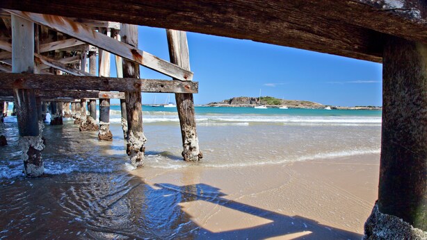 Un muelle de madera que se adentra en un mar azul cristalino, con una playa arenosa y afloramientos rocosos a lo lejos.