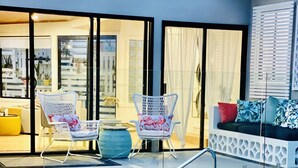Una piscina con borde de azulejos, un área de estar exterior blanca con cojines y una puerta de cristal que da a un espacio interior.