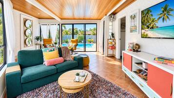 A living room with a green sofa, a coffee table, and a TV on a white wall. There is a pool visible through the glass doors.
