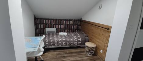 Une petite pièce agréable, dotée d’une tête de lit en bois, d’un lit recouvert d’une couverture à motifs, d’un bureau sur lequel est posée une carte et d’une bibliothèque remplie de livres.