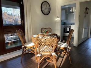 A dining area with a round table, four wicker chairs, a clock on the wall, and a view of a garden through a window. - One bedroom balcony apartment with allocated parking close to Gunwharf Quays (Portsmouth)
