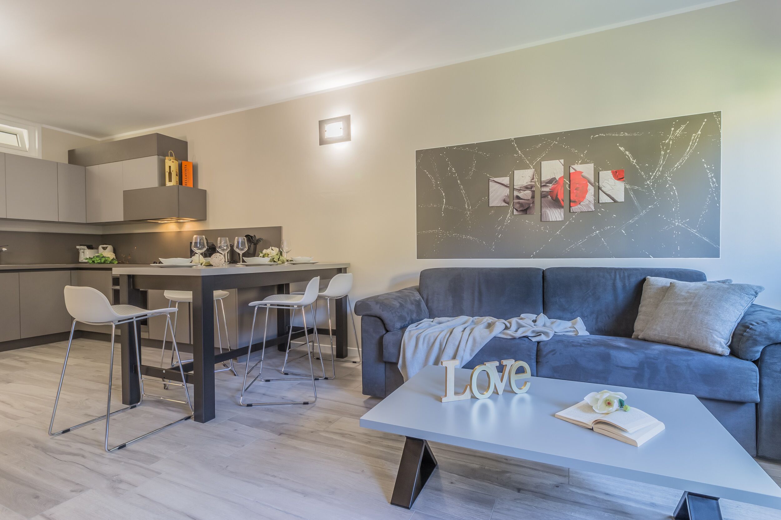 A modern living room with a grey sofa, a coffee table with a book and flowers, and a dining area with a table and chairs.