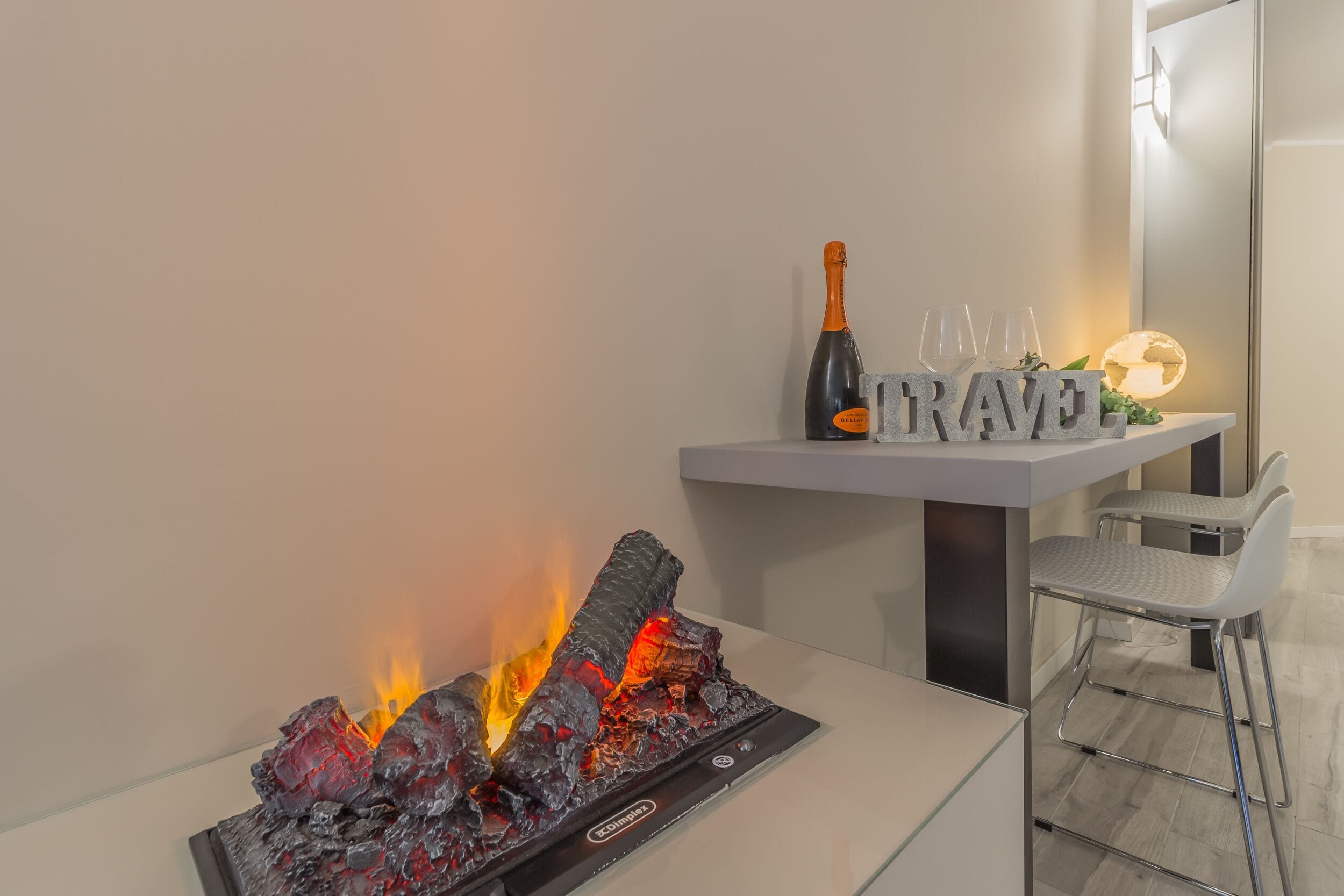 Une chambre moderne avec une table en verre, une cheminée où brûlent des bûches, une bouteille de champagne et un panneau indiquant « VOYAGE ».