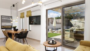 A modern living room with a dining area, large sliding glass doors to a backyard, and a flat-screen TV mounted on the wall. - Koin Villa Sunset,  With Heated Pool & Whirlpool (Perama, Rethymno)