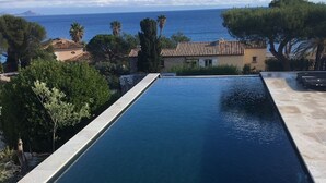 Uma piscina retangular com água azul cristalina, ladeada por ladrilhos de pedra e cercada por árvores e um edifício com telhado telhado.