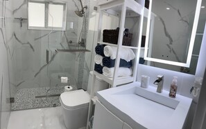 Une salle de bain moderne avec une douche à parois de verre, des murs en marbre, un lavabo blanc et des serviettes soigneusement pliées.