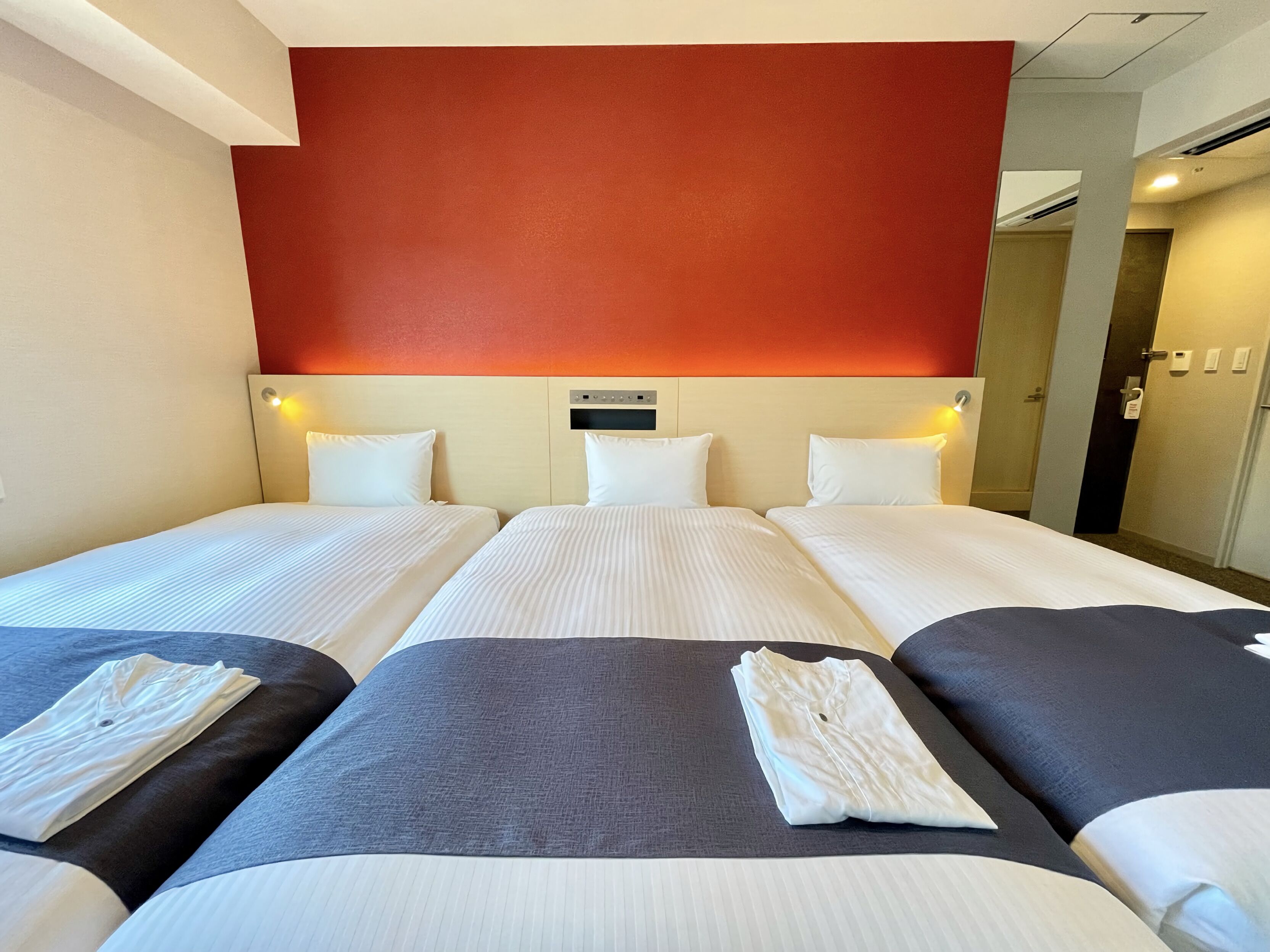 Ein Hotelzimmer mit drei Einzelbetten, jeweils mit weißer Bettwäsche und Kopfteil. Der Raum hat eine rote Akzentwand und ein modernes Design.