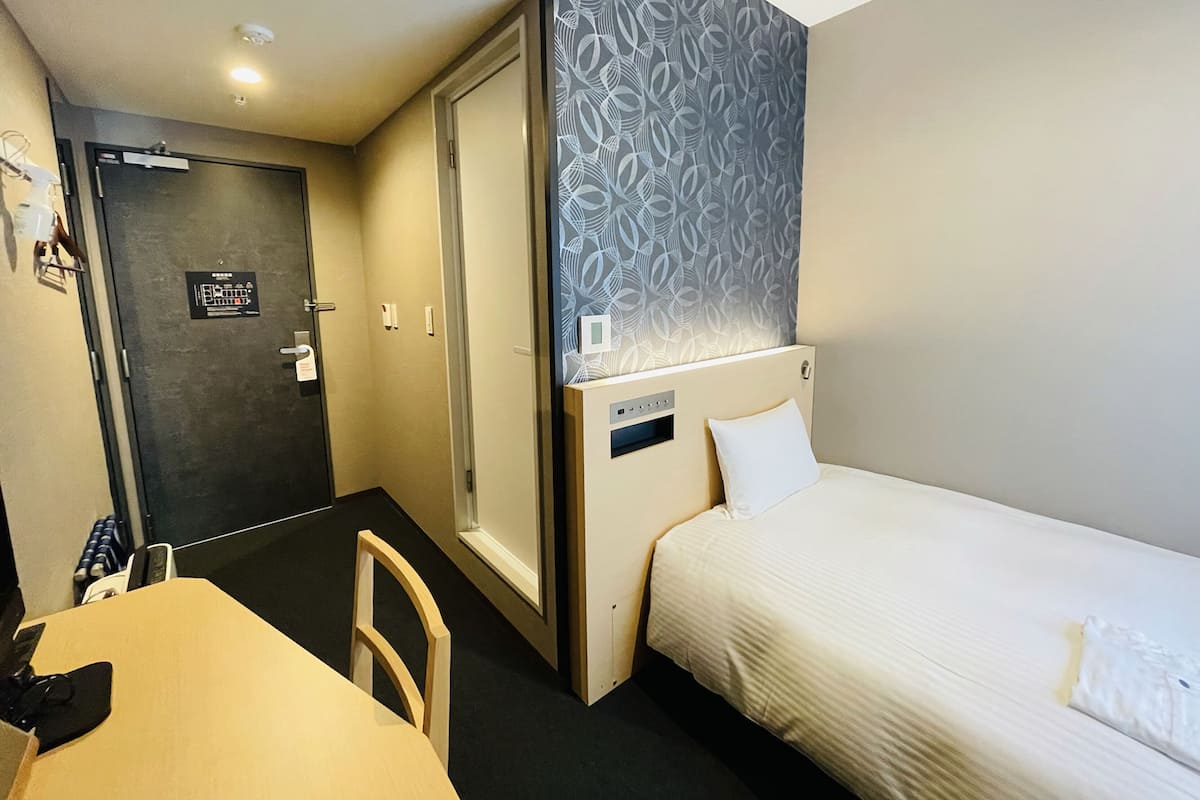 Een kleine hotelkamer met een enkel bed, een tafeltje en een stoel. Een deur leidt naar een badkamer, en de kamer heeft een behang met een patroon en een plafondlamp.
