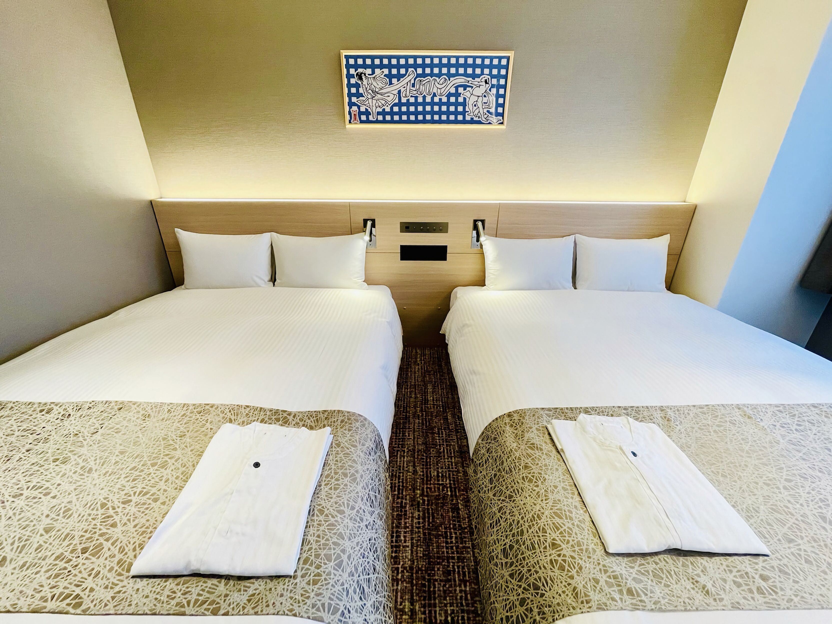 A hotel room with two beds, a headboard with a light switch, and a decorative wall piece above the beds.