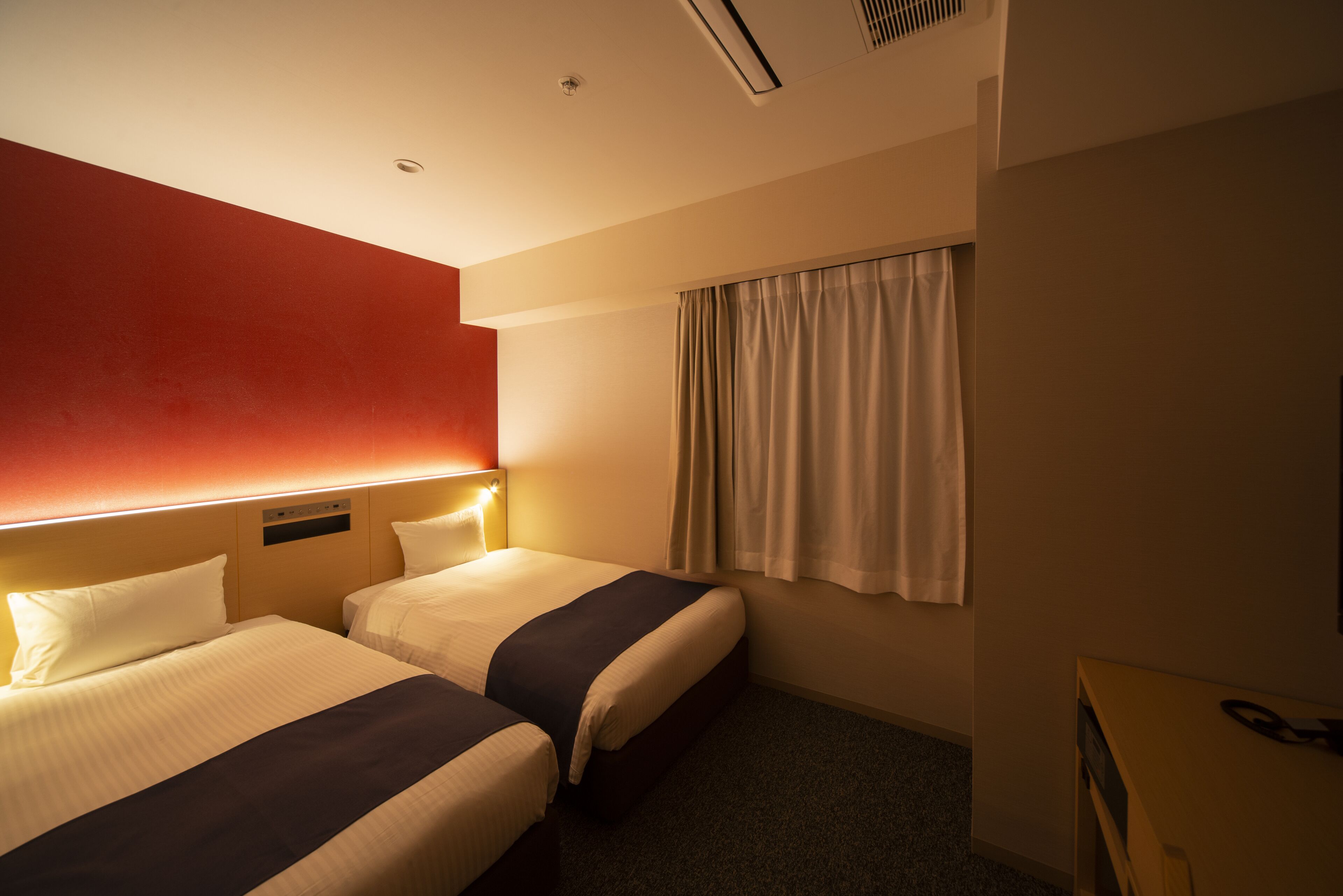 A hotel room with two beds, a red accent wall, a window with curtains, and a desk with a phone.