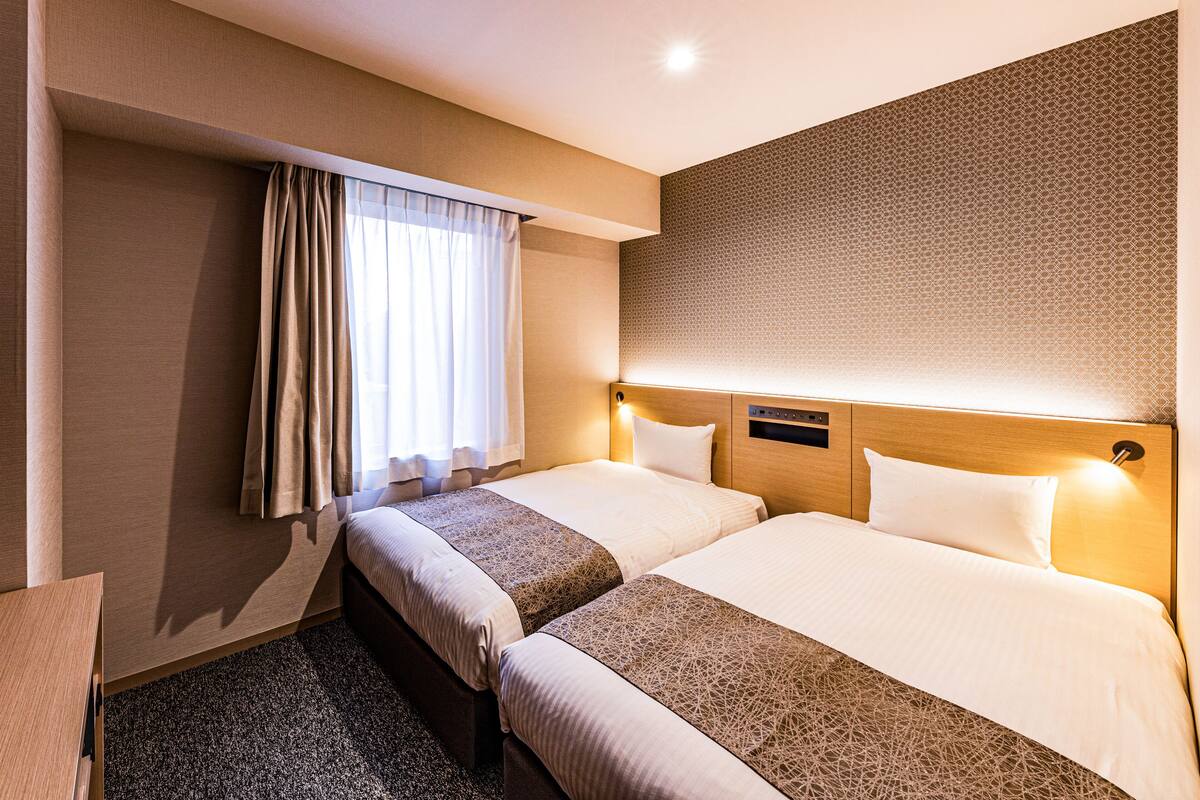 Een hotelkamer met twee bedden, een raam met gordijnen, een hoofdbord met lamp en een vloerbedekking.