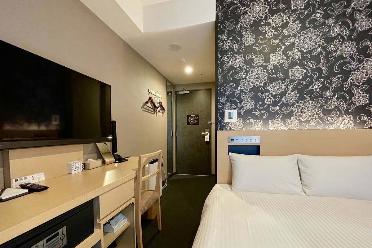 Hotelkamer met een bed, bureau met een flatscreen televisie, stoel, bureaulamp, klok en een wand met bloemenpatroon.