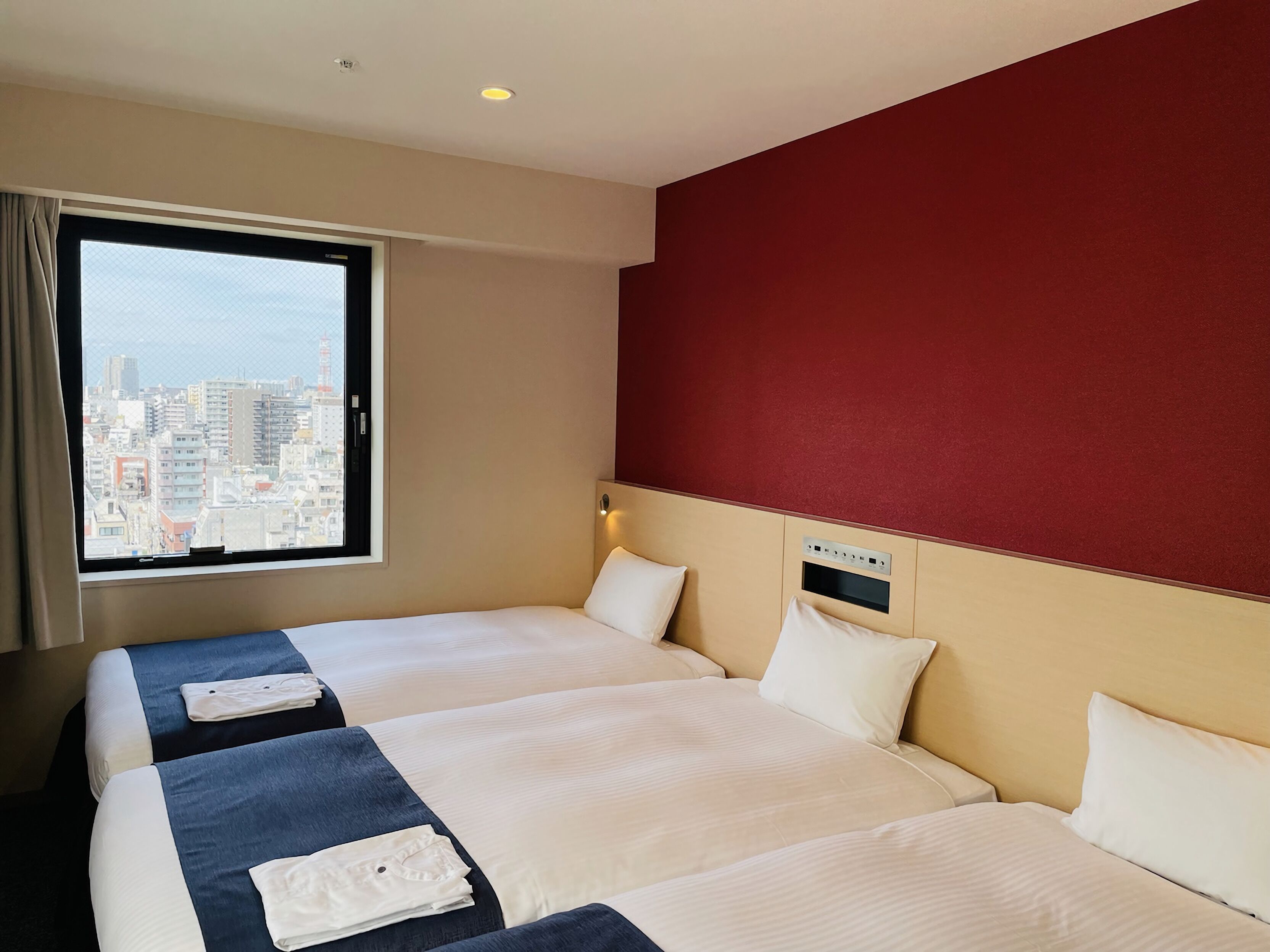 A hotel room with two beds, a large window with a city view, and a red accent wall.
