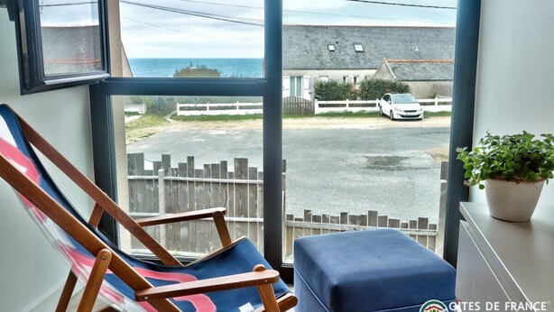 Une pièce dotée d’une grande fenêtre donnant sur un parking et un immeuble. On y trouve une chaise en bois avec un coussin coloré et un pouf bleu.