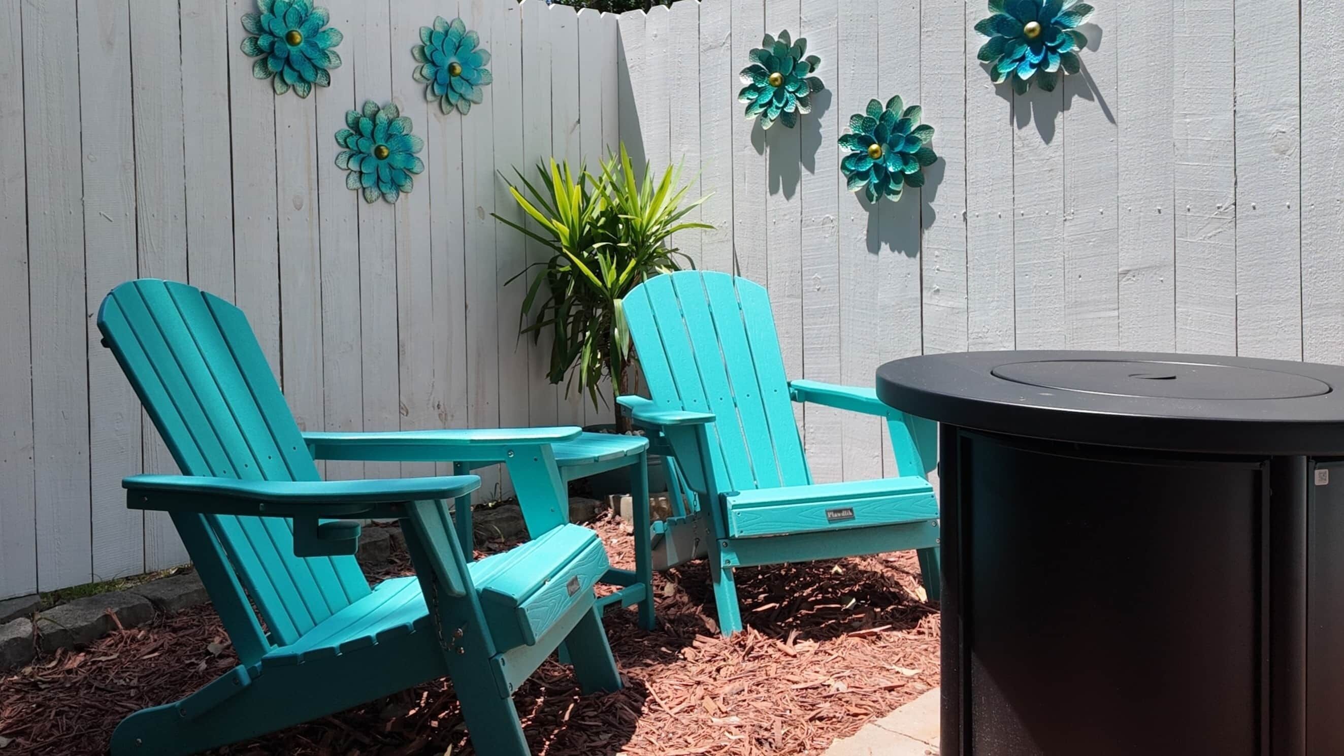 Deux chaises Adirondack turquoise, une petite table ronde et une plante en pot, le tout installé dans un coin du jardin, agrémenté d’une clôture blanche et de fleurs décoratives.