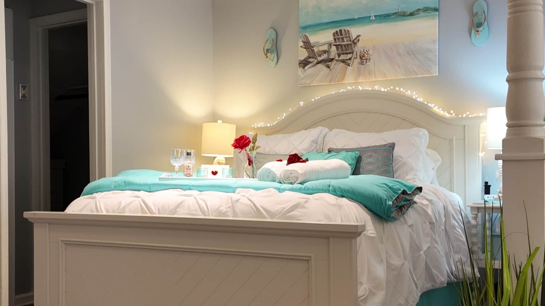A bedroom with a bed, bedside table, lamp, and a painting of a beach scene.