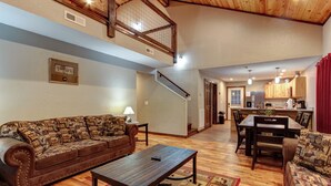 A spacious living room with a brown leather sofa, a wooden coffee table, and a patterned rug. A staircase leads to an upper level with a wooden railing. The kitchen is visible in the background with wooden cabinets and a red curtain. - Easy Access • No Steep Roads • Near Parkway • Hot Tub • Arcade • 3 Master Suites (Pigeon Forge)