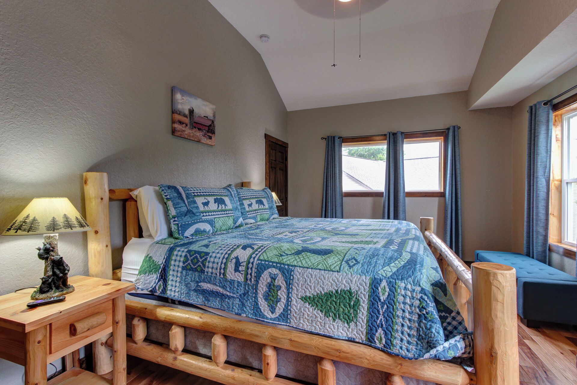 A bedroom with a wooden bed frame, a quilted bedspread, a nightstand with a lamp, and a window with curtains.