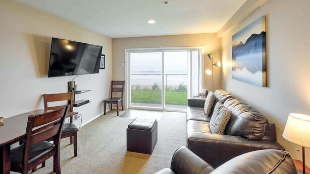 A living room with a flat-screen TV, a sofa, a dining table with chairs, and a large window with a view of greenery. - Ground Floor Beachfront Suite - Sleeps 4 - Pool and Sauna (Lincoln City)