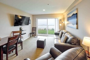 A living room with a flat-screen TV, a sofa, a dining table with chairs, and a large window with a view of greenery.