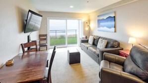 A living room with a wooden dining table, leather sofa, and a flat-screen TV mounted on the wall. - Ground Floor Beachfront Suite - Sleeps 4 - Pool and Sauna (Lincoln City)