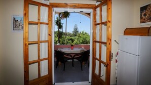 An open wooden door leading to a balcony with a table and chairs, a white refrigerator, and a view of greenery. - Alice & Teo Houses (Corfu)