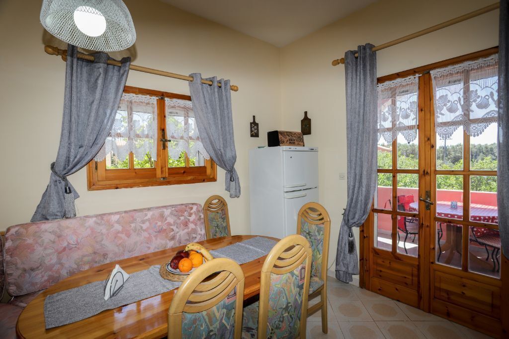 A dining area with a wooden table, chairs, and a bench. There is a window with curtains, a refrigerator, and a view of greenery outside.