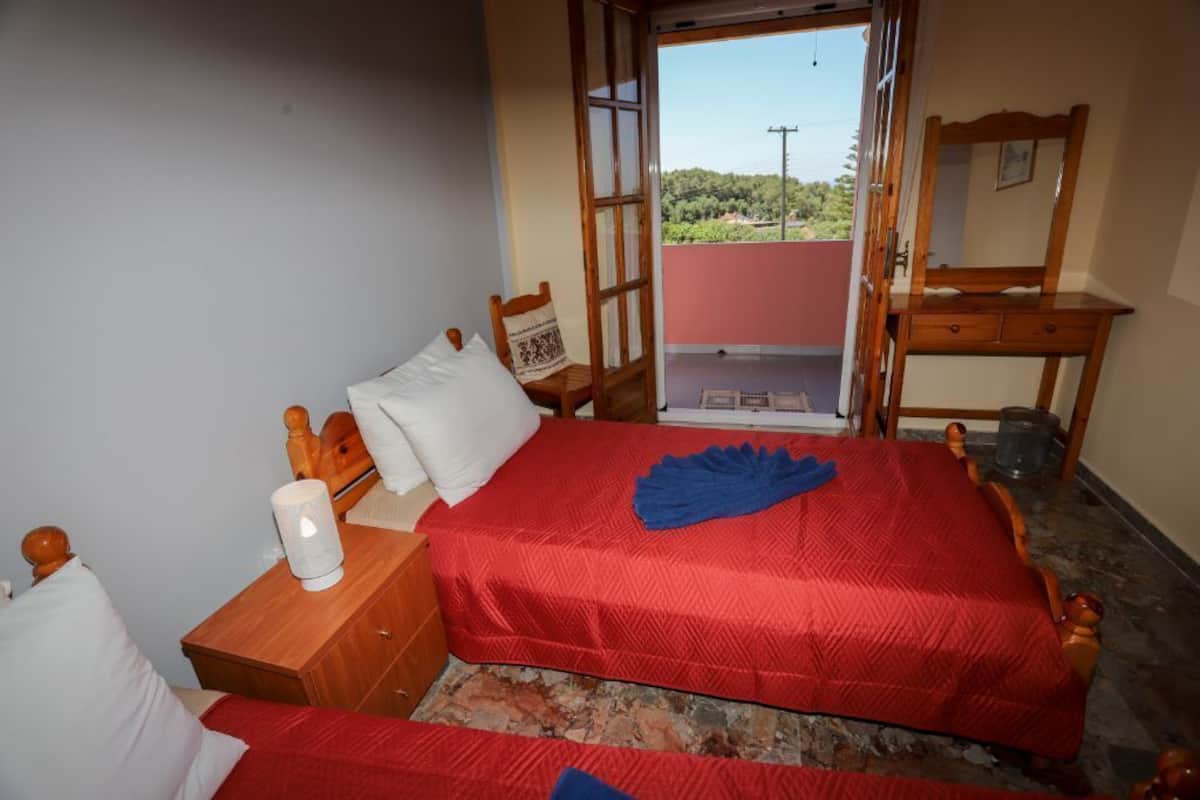 A bedroom with a red bedspread, a wooden chair, a bedside table with a lamp, and a view of greenery through an open door.