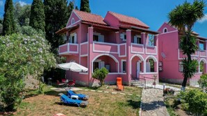 A pink villa with a tiled roof, a covered patio with lounge chairs, and a stone pathway leading to the entrance. - Alice & Teo Houses (Corfu)