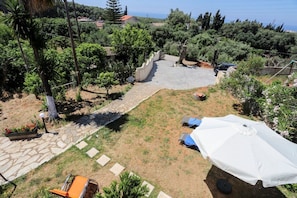 A backyard with a stone pathway, a small seating area, and a white umbrella. - Alice & Teo Houses (Corfu)