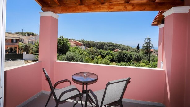 A balcony with a wooden ceiling, pink walls, and a view of a green landscape with houses. - Alice & Teo Houses (Corfu)