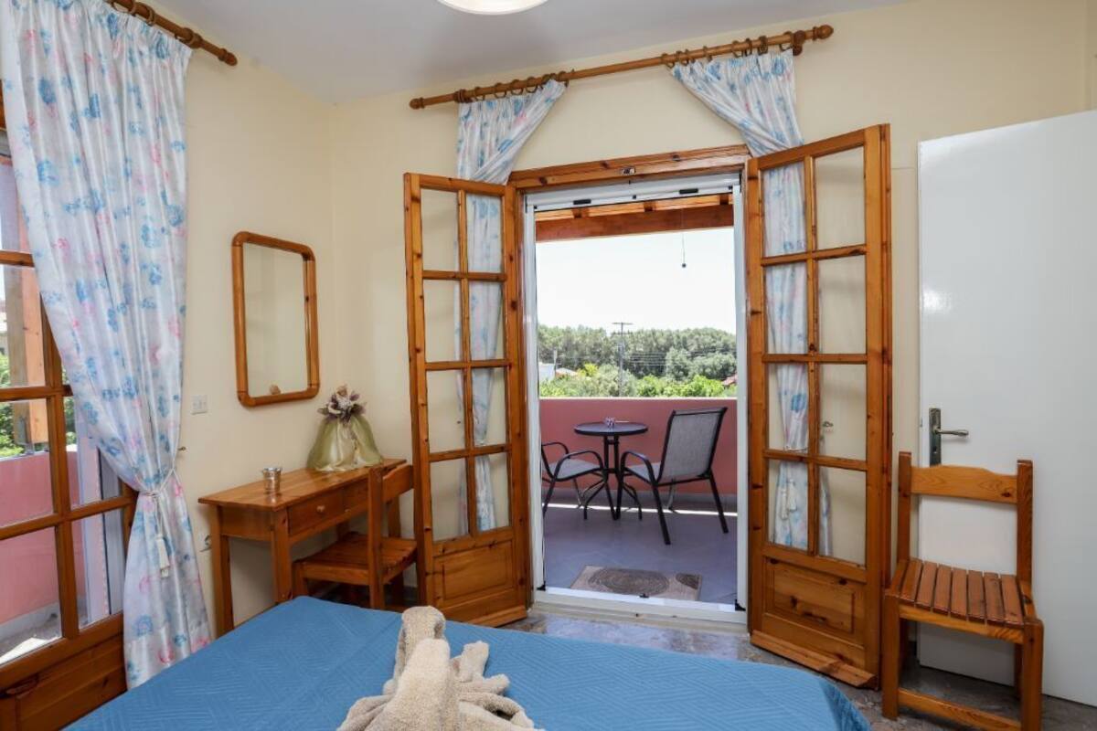 A bedroom with a bed, a wooden desk, a chair, a mirror, and an open door leading to a balcony with a table and chairs.