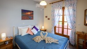 A bedroom with a bed, a nightstand, a lamp, a painting on the wall, and a window with curtains. - Alice & Teo Houses (Corfu)