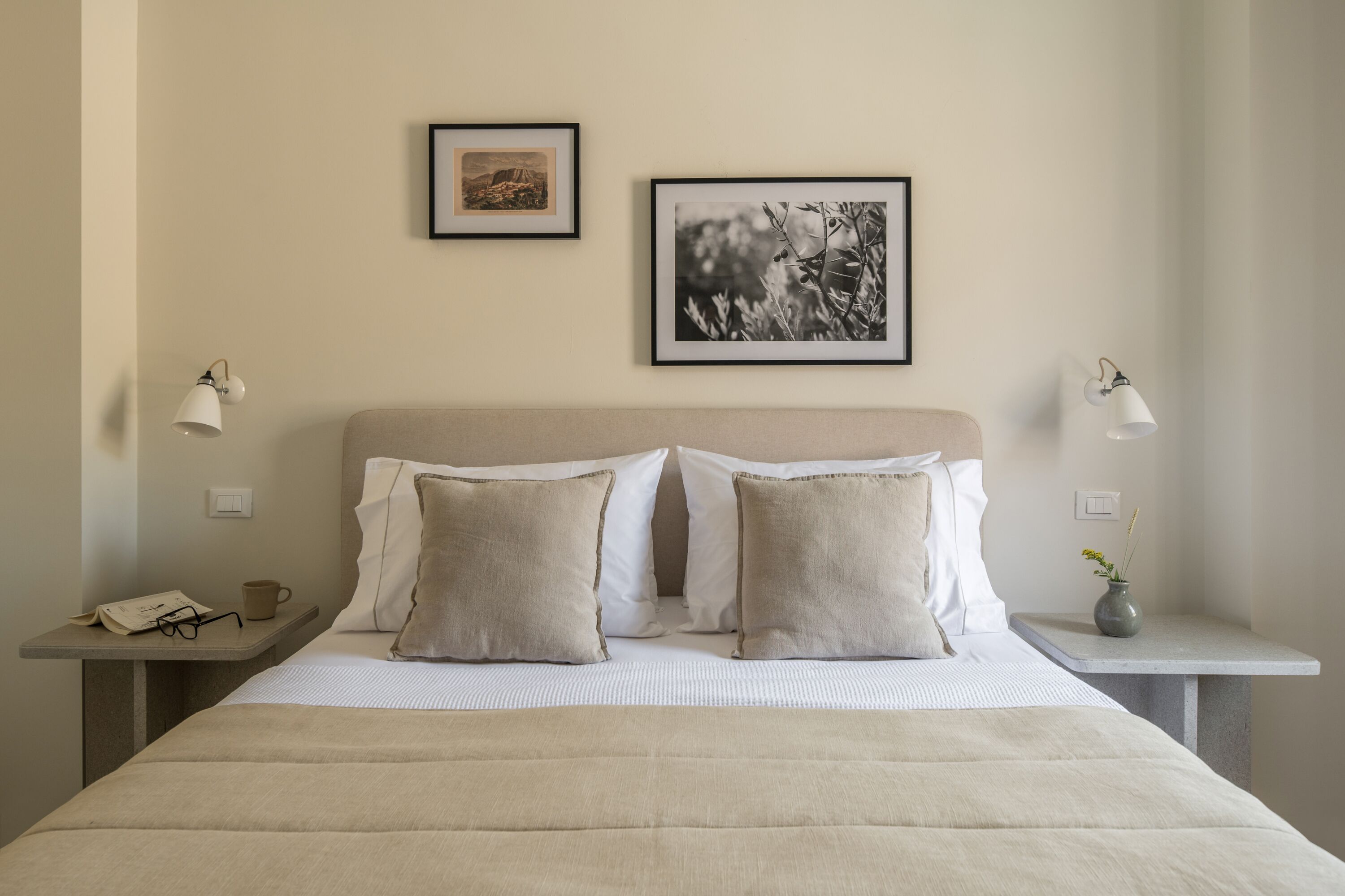 A neatly made bed with two pillows, a bedside table with a lamp, a small vase with flowers, and two framed pictures on the wall.