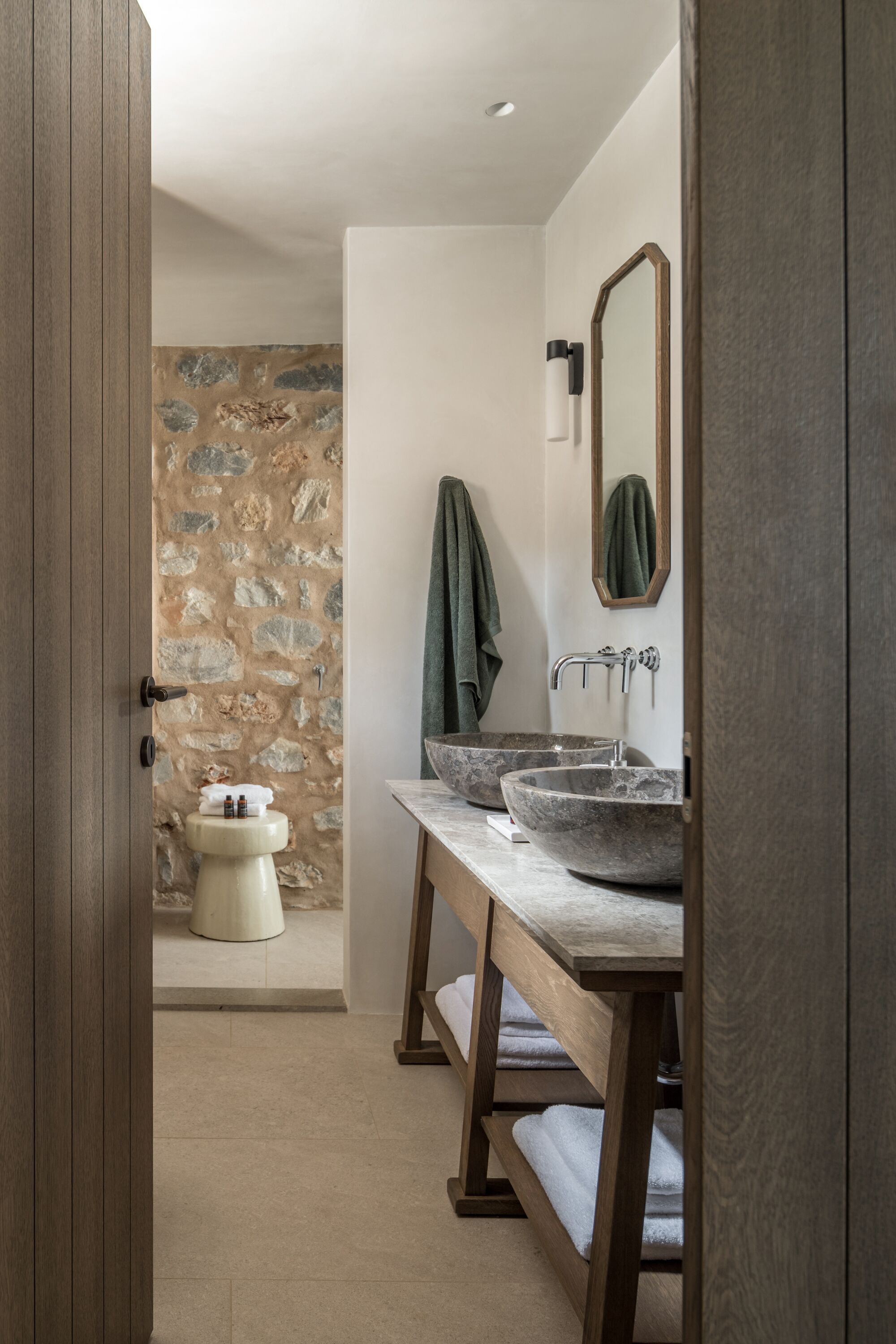 Un baño con pared de piedra, dos lavamanos, un espejo y un banco de madera.