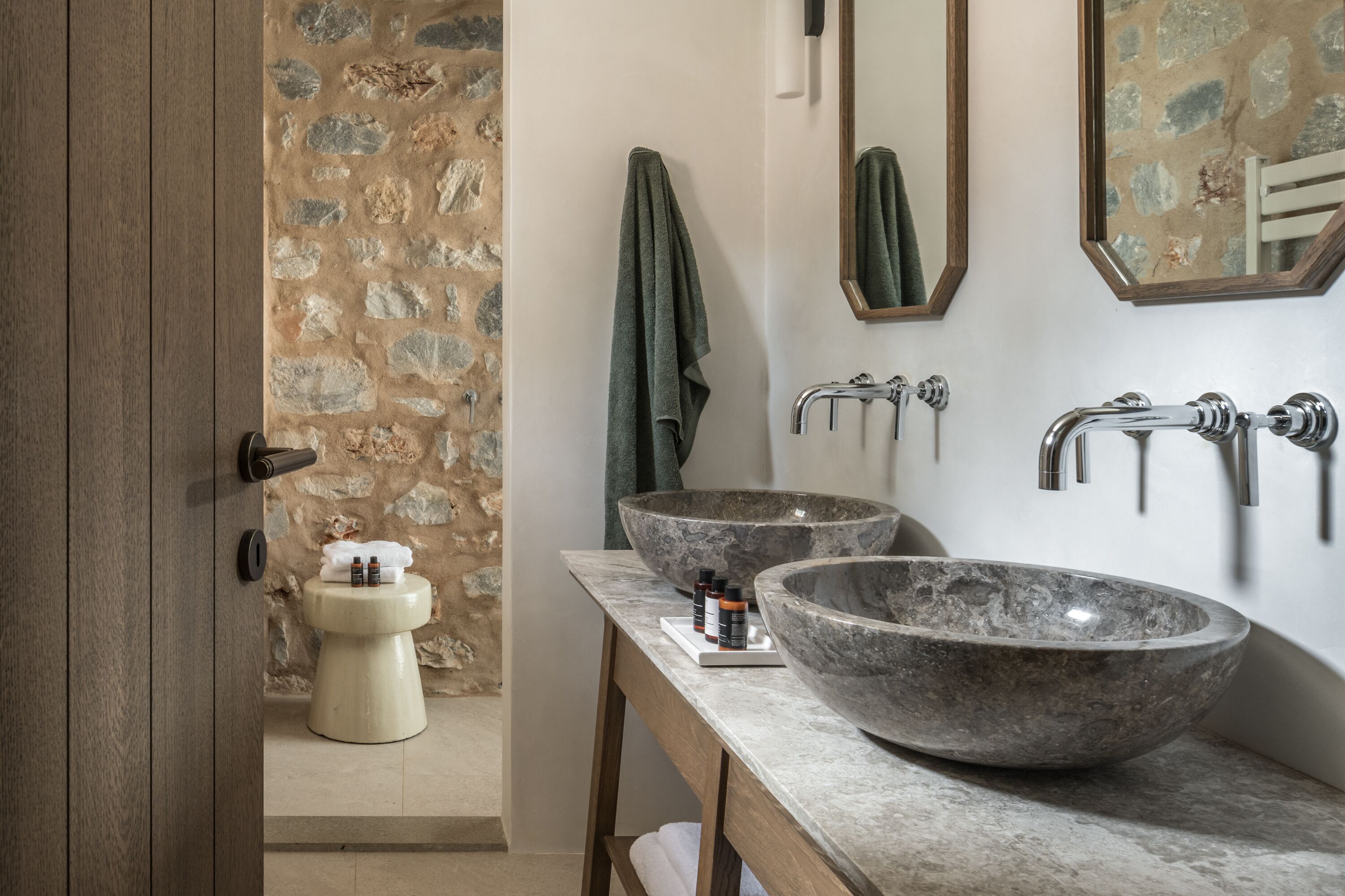 Ein Badezimmer mit Steinwand, Toilette, zwei großen Marmorwaschbecken und einem Holzhocker.