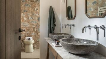 Ein Badezimmer mit Steinwand, Toilette, zwei großen Marmorwaschbecken und einem Holzhocker.