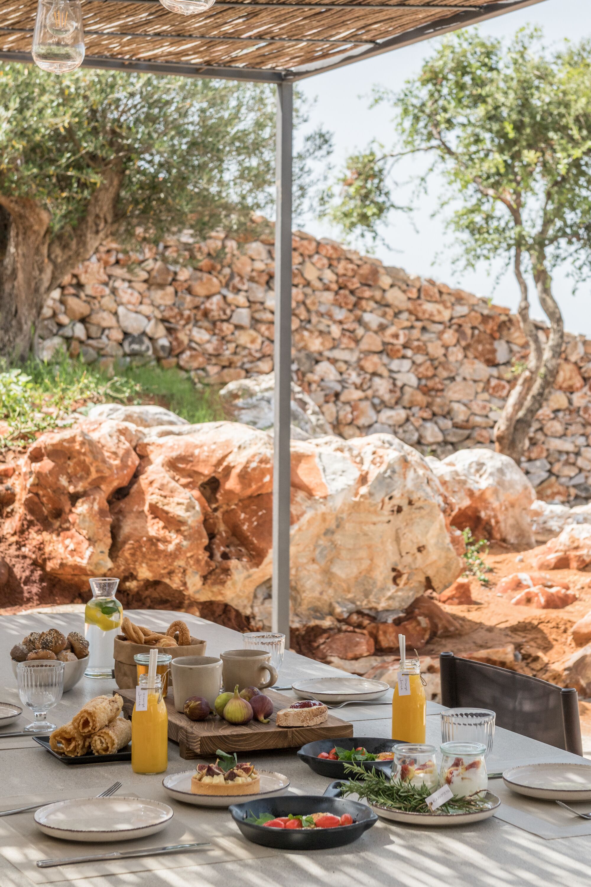 Un área de comedor al aire libre con un muro de piedra de fondo, una mesa dispuesta con alimentos para el desayuno y una vista despejada del paisaje circundante.